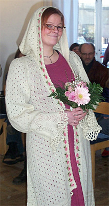 Frida visar brudkappan Julia. Den är stickad av tunnt ullgarn i hål och spetsmönster med rosa rosor i mönsterbård längs framkanterna. design textilkonstnär katrin bawah.